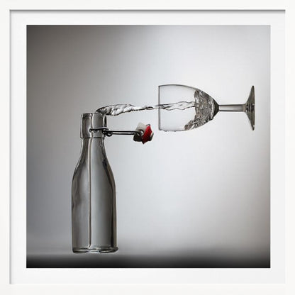 A conceptual photograph in a light wood frame depicting water pouring horizontally from an open swing-top glass bottle into a floating, sideways wine glass against a simple gray background, capturing the liquid in mid-air. Print