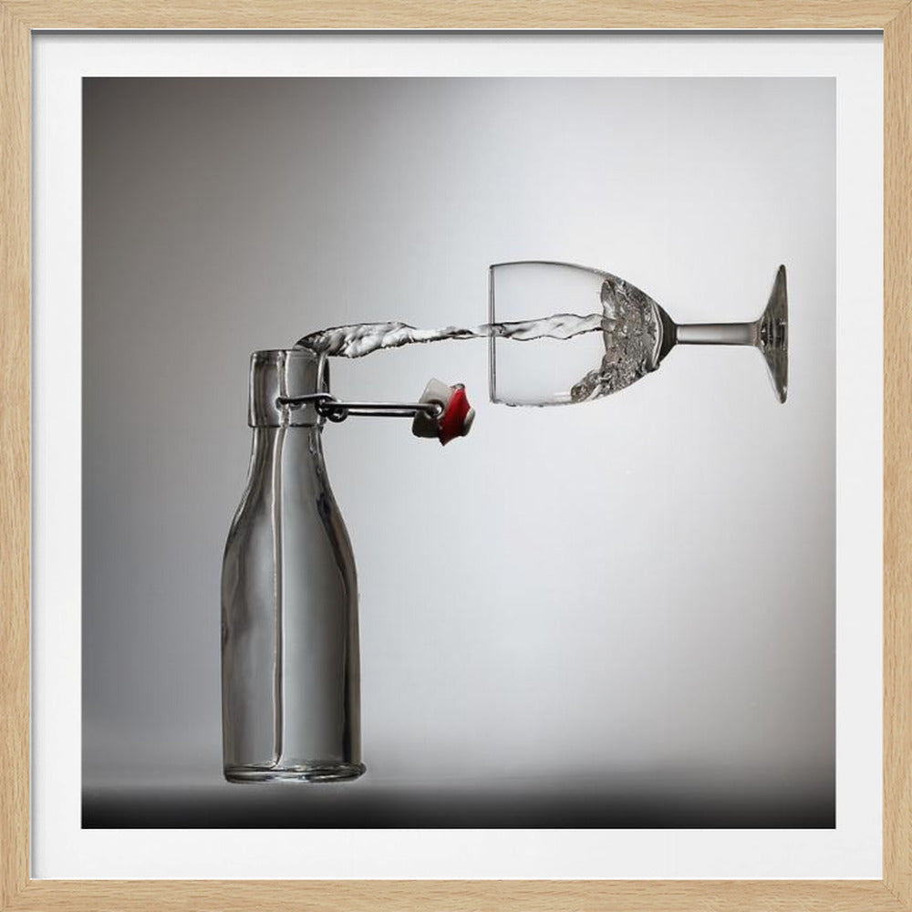 A conceptual photograph in a light wood frame depicting water pouring horizontally from an open swing-top glass bottle into a floating, sideways wine glass against a simple gray background, capturing the liquid in mid-air. Print