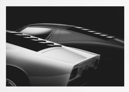 A dramatic black and white studio photograph showcasing the sleek, curved lines of two classic sports cars, with a white car in the foreground and a dark one in the background, set against a black backdrop. Wall Art