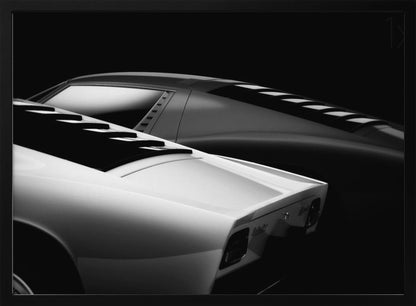A dramatic black and white studio photograph showcasing the sleek, curved lines of two classic sports cars, with a white car in the foreground and a dark one in the background, set against a black backdrop. Wall Art