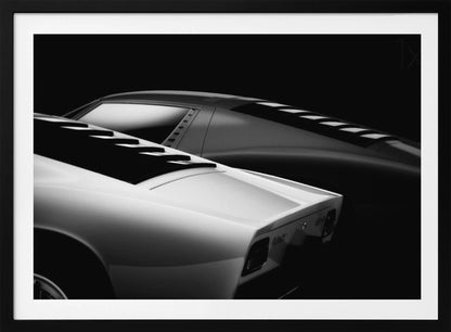 A dramatic black and white studio photograph showcasing the sleek, curved lines of two classic sports cars, with a white car in the foreground and a dark one in the background, set against a black backdrop. Wall Art