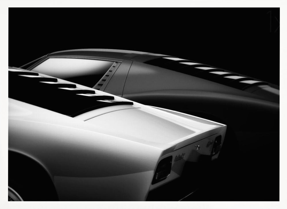 A dramatic black and white studio photograph showcasing the sleek, curved lines of two classic sports cars, with a white car in the foreground and a dark one in the background, set against a black backdrop. Wall Art