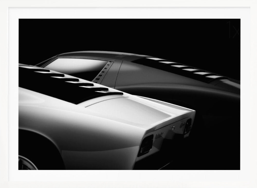 A dramatic black and white studio photograph showcasing the sleek, curved lines of two classic sports cars, with a white car in the foreground and a dark one in the background, set against a black backdrop. Wall Art