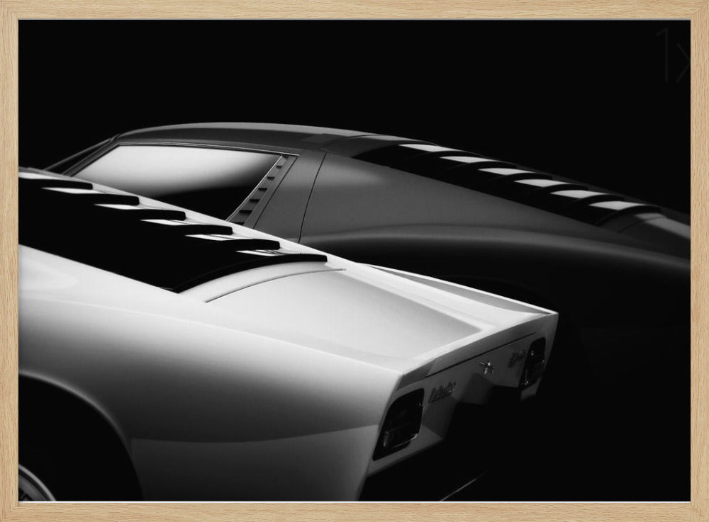 A dramatic black and white studio photograph showcasing the sleek, curved lines of two classic sports cars, with a white car in the foreground and a dark one in the background, set against a black backdrop. Wall Art