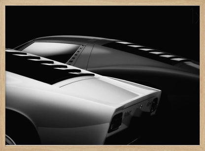 A dramatic black and white studio photograph showcasing the sleek, curved lines of two classic sports cars, with a white car in the foreground and a dark one in the background, set against a black backdrop. Wall Art