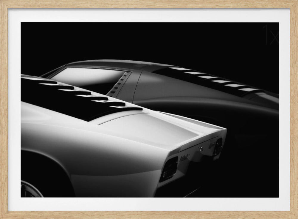 A dramatic black and white studio photograph showcasing the sleek, curved lines of two classic sports cars, with a white car in the foreground and a dark one in the background, set against a black backdrop. Wall Art
