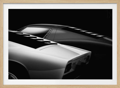 A dramatic black and white studio photograph showcasing the sleek, curved lines of two classic sports cars, with a white car in the foreground and a dark one in the background, set against a black backdrop. Wall Art