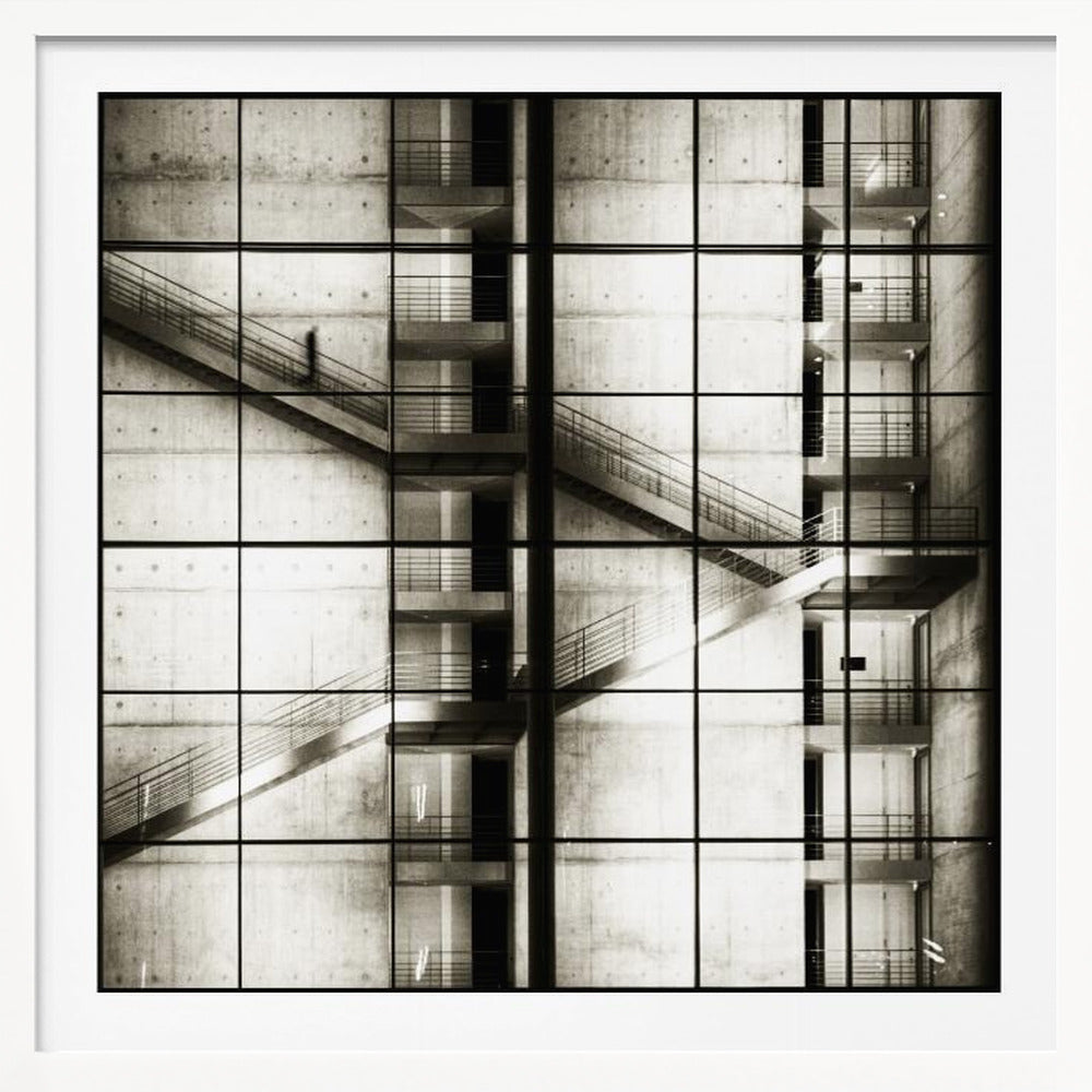 A framed black and white photograph of a modern concrete building's interior, seen through a grid of window panes. The image captures multiple levels of staircases and walkways crisscrossing in a complex, geometric pattern. A shadowy, blurred figure of a person can be seen walking up a staircase on the upper left. Wall Art