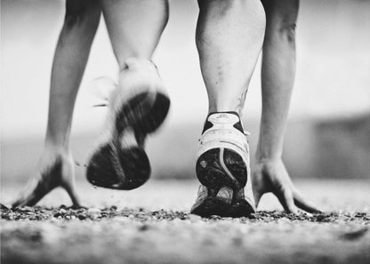 A low-angle, black and white action shot of a runner's legs and hands at the starting position on a gravel track. One foot is planted firmly while the other is in motion, blurred as they push off to start a race. Decor