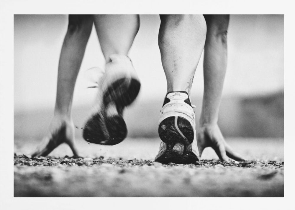 A low-angle, black and white action shot of a runner's legs and hands at the starting position on a gravel track. One foot is planted firmly while the other is in motion, blurred as they push off to start a race. Decor