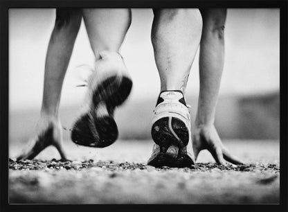 A low-angle, black and white action shot of a runner's legs and hands at the starting position on a gravel track. One foot is planted firmly while the other is in motion, blurred as they push off to start a race. Decor