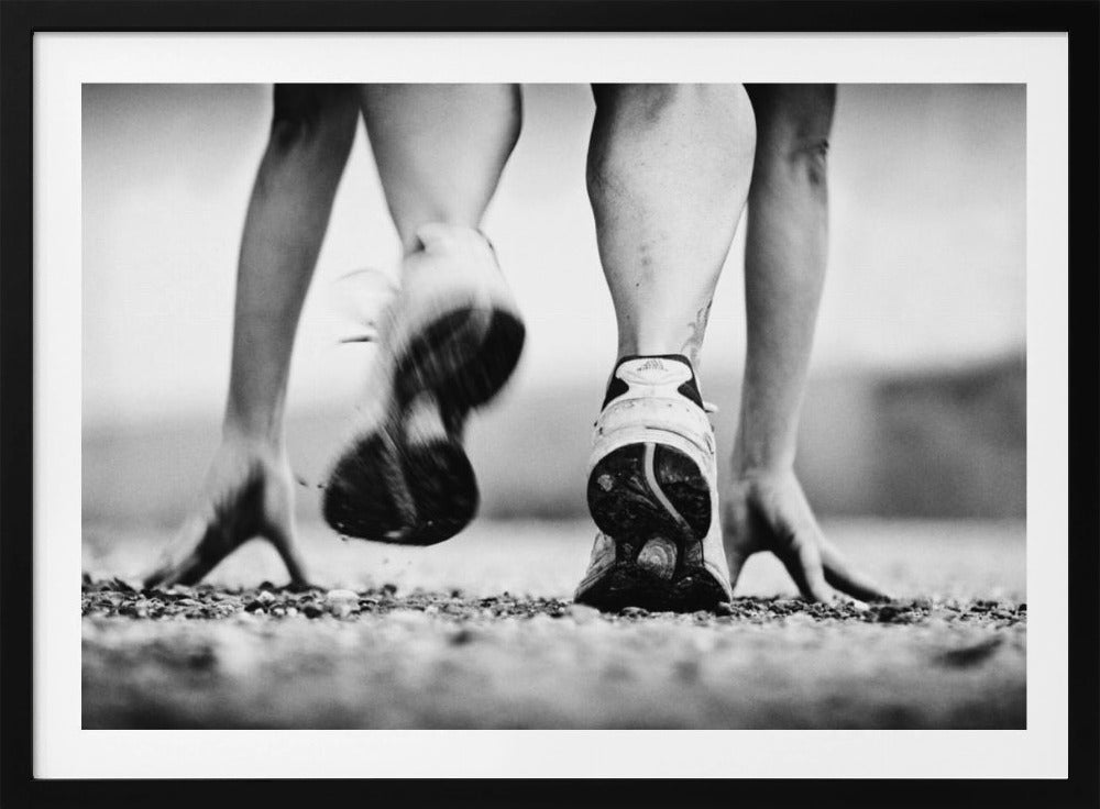 A low-angle, black and white action shot of a runner's legs and hands at the starting position on a gravel track. One foot is planted firmly while the other is in motion, blurred as they push off to start a race. Decor