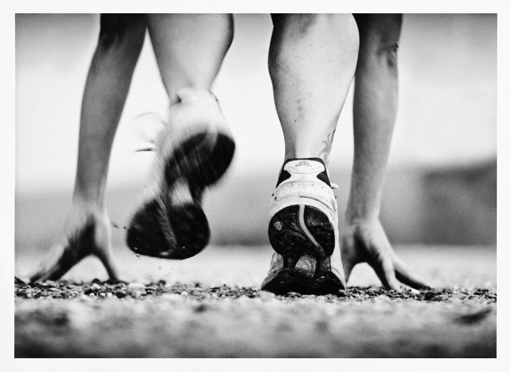 A low-angle, black and white action shot of a runner's legs and hands at the starting position on a gravel track. One foot is planted firmly while the other is in motion, blurred as they push off to start a race. Decor