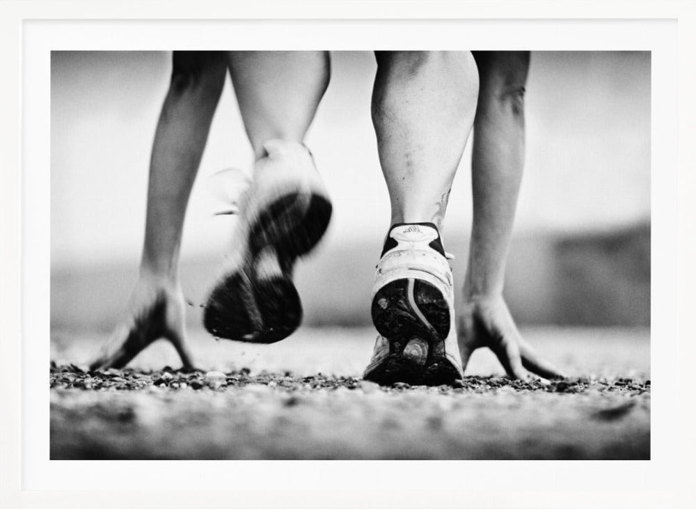 A low-angle, black and white action shot of a runner's legs and hands at the starting position on a gravel track. One foot is planted firmly while the other is in motion, blurred as they push off to start a race. Decor