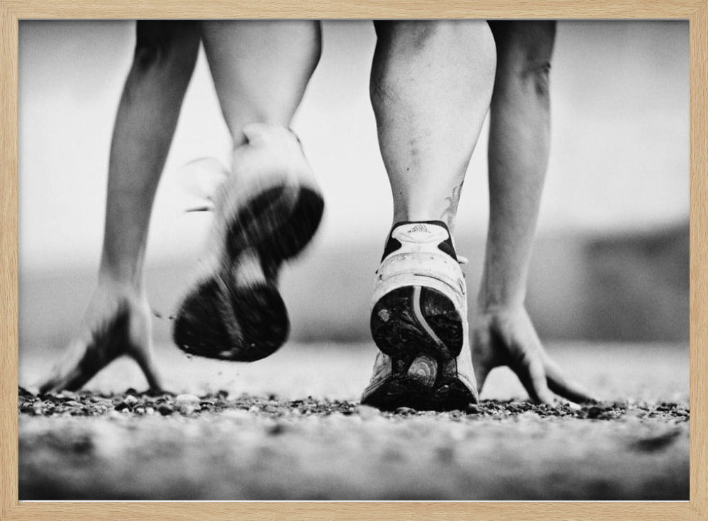 A low-angle, black and white action shot of a runner's legs and hands at the starting position on a gravel track. One foot is planted firmly while the other is in motion, blurred as they push off to start a race. Decor