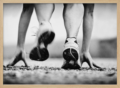 A low-angle, black and white action shot of a runner's legs and hands at the starting position on a gravel track. One foot is planted firmly while the other is in motion, blurred as they push off to start a race. Decor