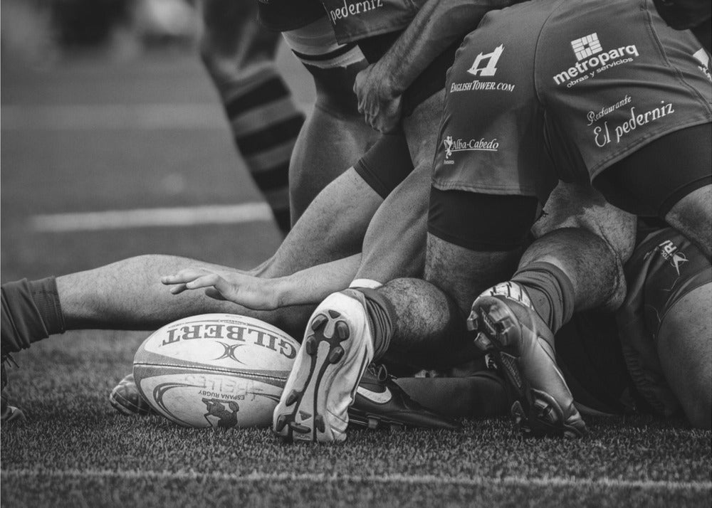 A dynamic black and white, low-angle photograph capturing the intensity of a rugby ruck. A Gilbert rugby ball sits on the turf in the foreground, surrounded by a tangle of players' muscular legs, arms, and cleats, conveying the raw power and action of the sport. Wall Art
