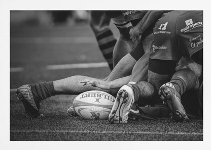 A dynamic black and white, low-angle photograph capturing the intensity of a rugby ruck. A Gilbert rugby ball sits on the turf in the foreground, surrounded by a tangle of players' muscular legs, arms, and cleats, conveying the raw power and action of the sport. Wall Art
