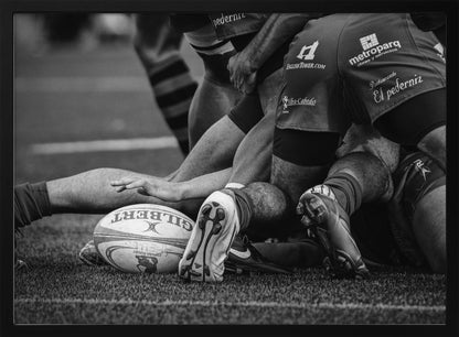 A dynamic black and white, low-angle photograph capturing the intensity of a rugby ruck. A Gilbert rugby ball sits on the turf in the foreground, surrounded by a tangle of players' muscular legs, arms, and cleats, conveying the raw power and action of the sport. Wall Art