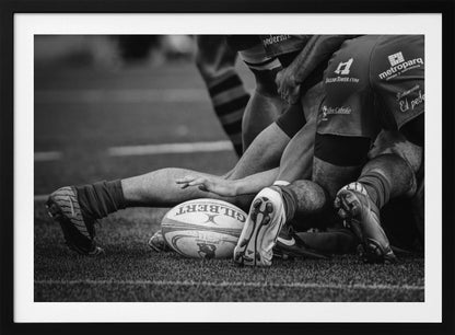 A dynamic black and white, low-angle photograph capturing the intensity of a rugby ruck. A Gilbert rugby ball sits on the turf in the foreground, surrounded by a tangle of players' muscular legs, arms, and cleats, conveying the raw power and action of the sport. Wall Art
