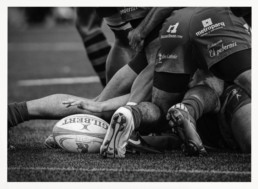 A dynamic black and white, low-angle photograph capturing the intensity of a rugby ruck. A Gilbert rugby ball sits on the turf in the foreground, surrounded by a tangle of players' muscular legs, arms, and cleats, conveying the raw power and action of the sport. Wall Art