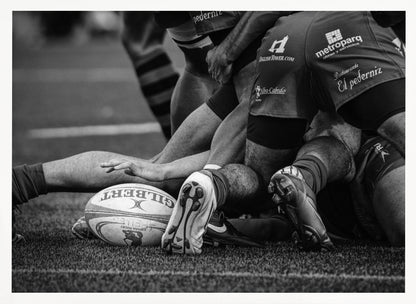 A dynamic black and white, low-angle photograph capturing the intensity of a rugby ruck. A Gilbert rugby ball sits on the turf in the foreground, surrounded by a tangle of players' muscular legs, arms, and cleats, conveying the raw power and action of the sport. Wall Art