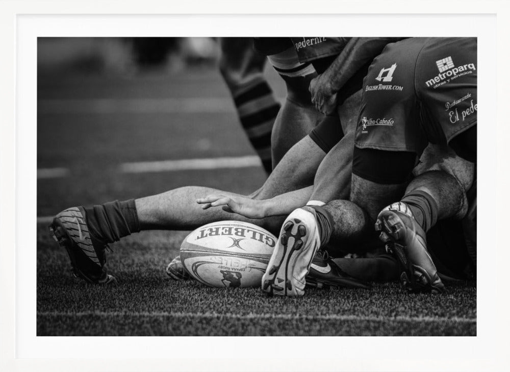 A dynamic black and white, low-angle photograph capturing the intensity of a rugby ruck. A Gilbert rugby ball sits on the turf in the foreground, surrounded by a tangle of players' muscular legs, arms, and cleats, conveying the raw power and action of the sport. Wall Art