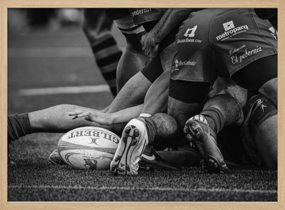 A dynamic black and white, low-angle photograph capturing the intensity of a rugby ruck. A Gilbert rugby ball sits on the turf in the foreground, surrounded by a tangle of players' muscular legs, arms, and cleats, conveying the raw power and action of the sport. Wall Art