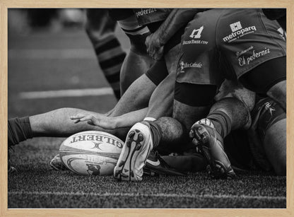 A dynamic black and white, low-angle photograph capturing the intensity of a rugby ruck. A Gilbert rugby ball sits on the turf in the foreground, surrounded by a tangle of players' muscular legs, arms, and cleats, conveying the raw power and action of the sport. Wall Art