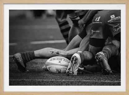 A dynamic black and white, low-angle photograph capturing the intensity of a rugby ruck. A Gilbert rugby ball sits on the turf in the foreground, surrounded by a tangle of players' muscular legs, arms, and cleats, conveying the raw power and action of the sport. Wall Art