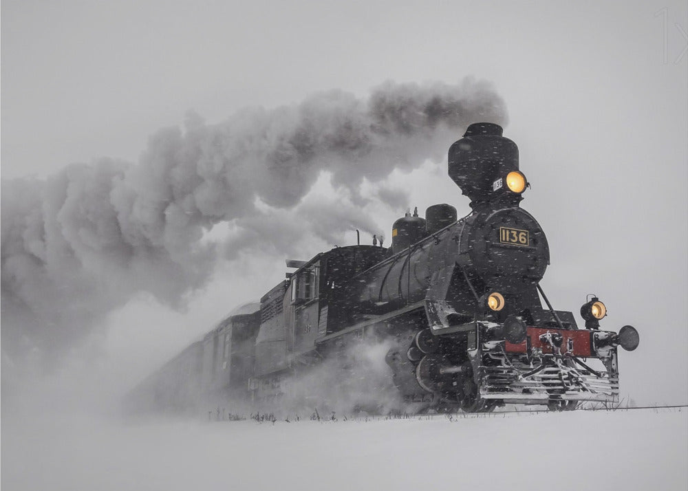 A dramatic black and white photograph of a classic steam train, number 1136, plowing through a heavy snowstorm, with its headlights glowing and a large plume of smoke trailing behind it. The image is presented within a silver frame. Print
