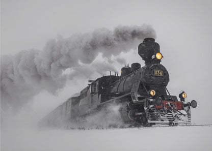 A dramatic black and white photograph of a classic steam train, number 1136, plowing through a heavy snowstorm, with its headlights glowing and a large plume of smoke trailing behind it. The image is presented within a silver frame. Print