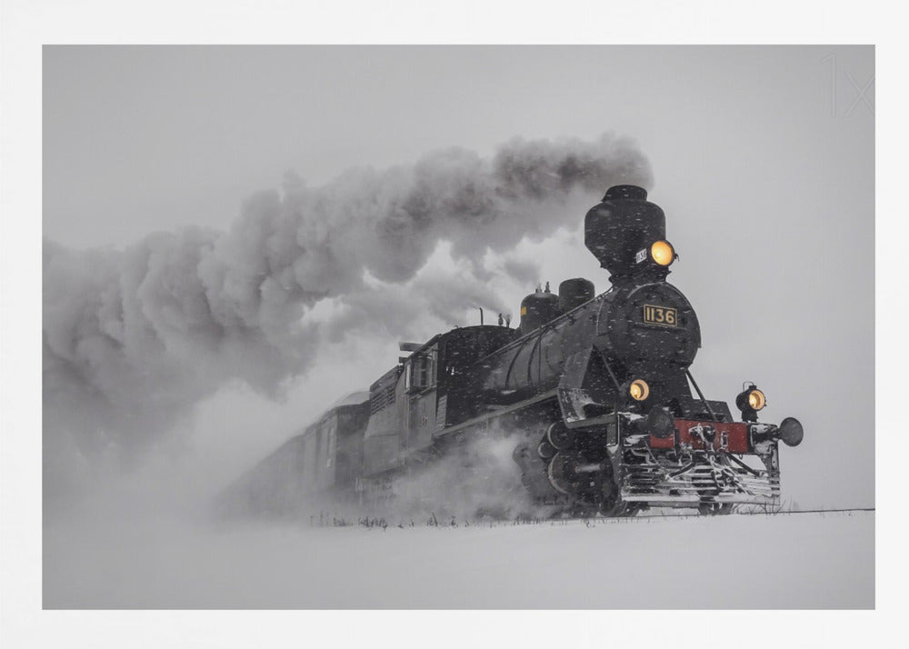 A dramatic black and white photograph of a classic steam train, number 1136, plowing through a heavy snowstorm, with its headlights glowing and a large plume of smoke trailing behind it. The image is presented within a silver frame. Print