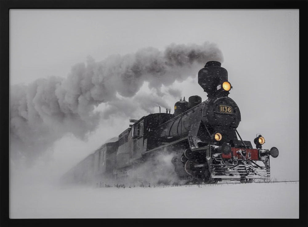 A dramatic black and white photograph of a classic steam train, number 1136, plowing through a heavy snowstorm, with its headlights glowing and a large plume of smoke trailing behind it. The image is presented within a silver frame. Print