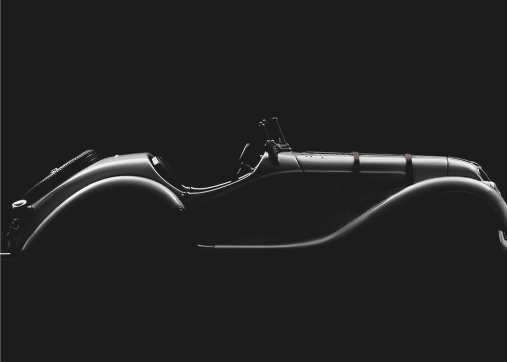 A dramatic black and white photograph showcasing the elegant silhouette of a vintage roadster. The side profile of the classic car is highlighted by subtle light, emphasizing its long hood and sweeping fenders against a solid black background, all within a brushed silver frame. Print