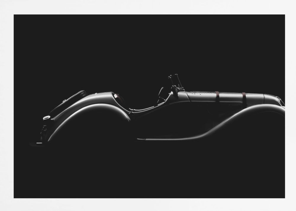 A dramatic black and white photograph showcasing the elegant silhouette of a vintage roadster. The side profile of the classic car is highlighted by subtle light, emphasizing its long hood and sweeping fenders against a solid black background, all within a brushed silver frame. Print