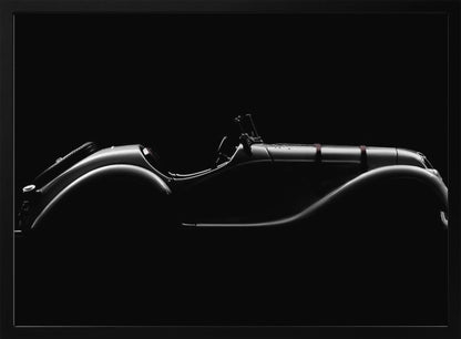 A dramatic black and white photograph showcasing the elegant silhouette of a vintage roadster. The side profile of the classic car is highlighted by subtle light, emphasizing its long hood and sweeping fenders against a solid black background, all within a brushed silver frame. Print