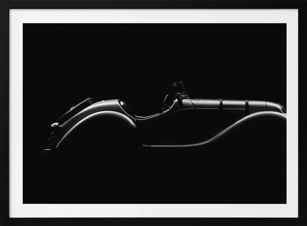 A dramatic black and white photograph showcasing the elegant silhouette of a vintage roadster. The side profile of the classic car is highlighted by subtle light, emphasizing its long hood and sweeping fenders against a solid black background, all within a brushed silver frame. Print