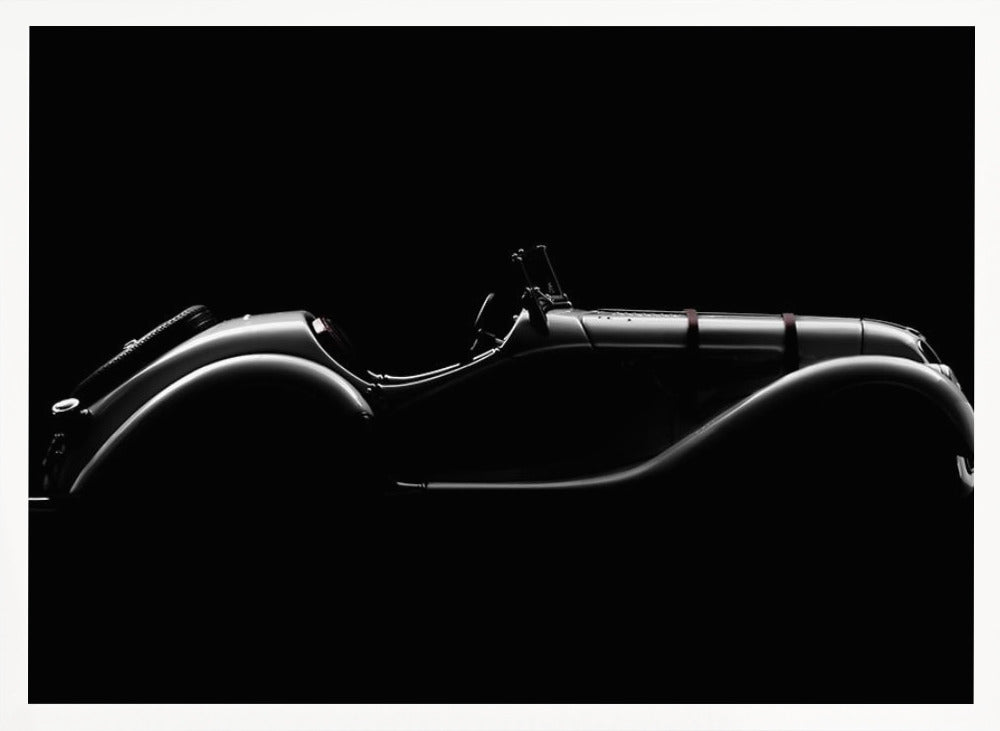A dramatic black and white photograph showcasing the elegant silhouette of a vintage roadster. The side profile of the classic car is highlighted by subtle light, emphasizing its long hood and sweeping fenders against a solid black background, all within a brushed silver frame. Print