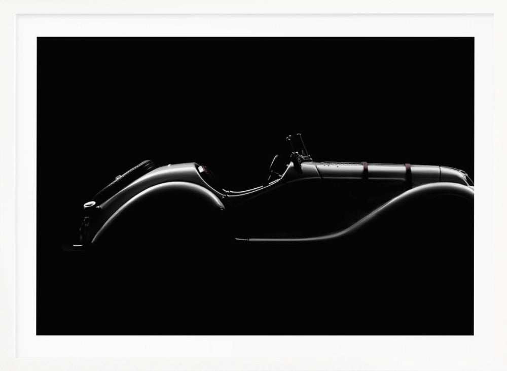 A dramatic black and white photograph showcasing the elegant silhouette of a vintage roadster. The side profile of the classic car is highlighted by subtle light, emphasizing its long hood and sweeping fenders against a solid black background, all within a brushed silver frame. Print