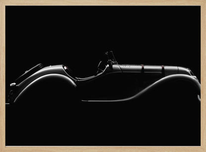 A dramatic black and white photograph showcasing the elegant silhouette of a vintage roadster. The side profile of the classic car is highlighted by subtle light, emphasizing its long hood and sweeping fenders against a solid black background, all within a brushed silver frame. Print