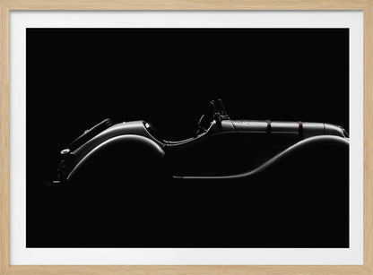 A dramatic black and white photograph showcasing the elegant silhouette of a vintage roadster. The side profile of the classic car is highlighted by subtle light, emphasizing its long hood and sweeping fenders against a solid black background, all within a brushed silver frame. Print