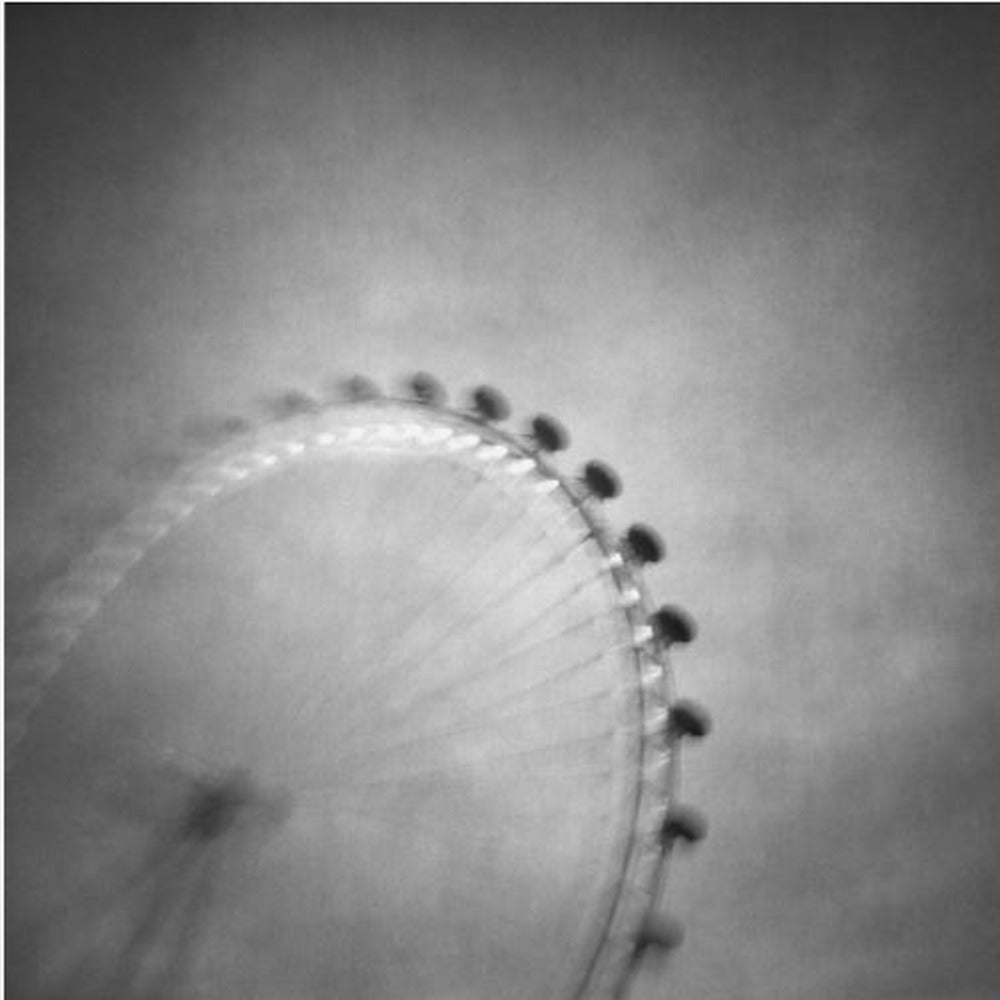A moody, black and white long-exposure photograph of a large Ferris wheel, creating a soft, blurry effect of its rotation against a grainy gray sky. The artwork is presented in a simple, light wood frame. Poster