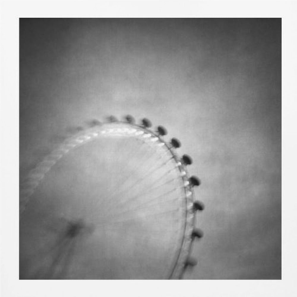 A moody, black and white long-exposure photograph of a large Ferris wheel, creating a soft, blurry effect of its rotation against a grainy gray sky. The artwork is presented in a simple, light wood frame. Poster