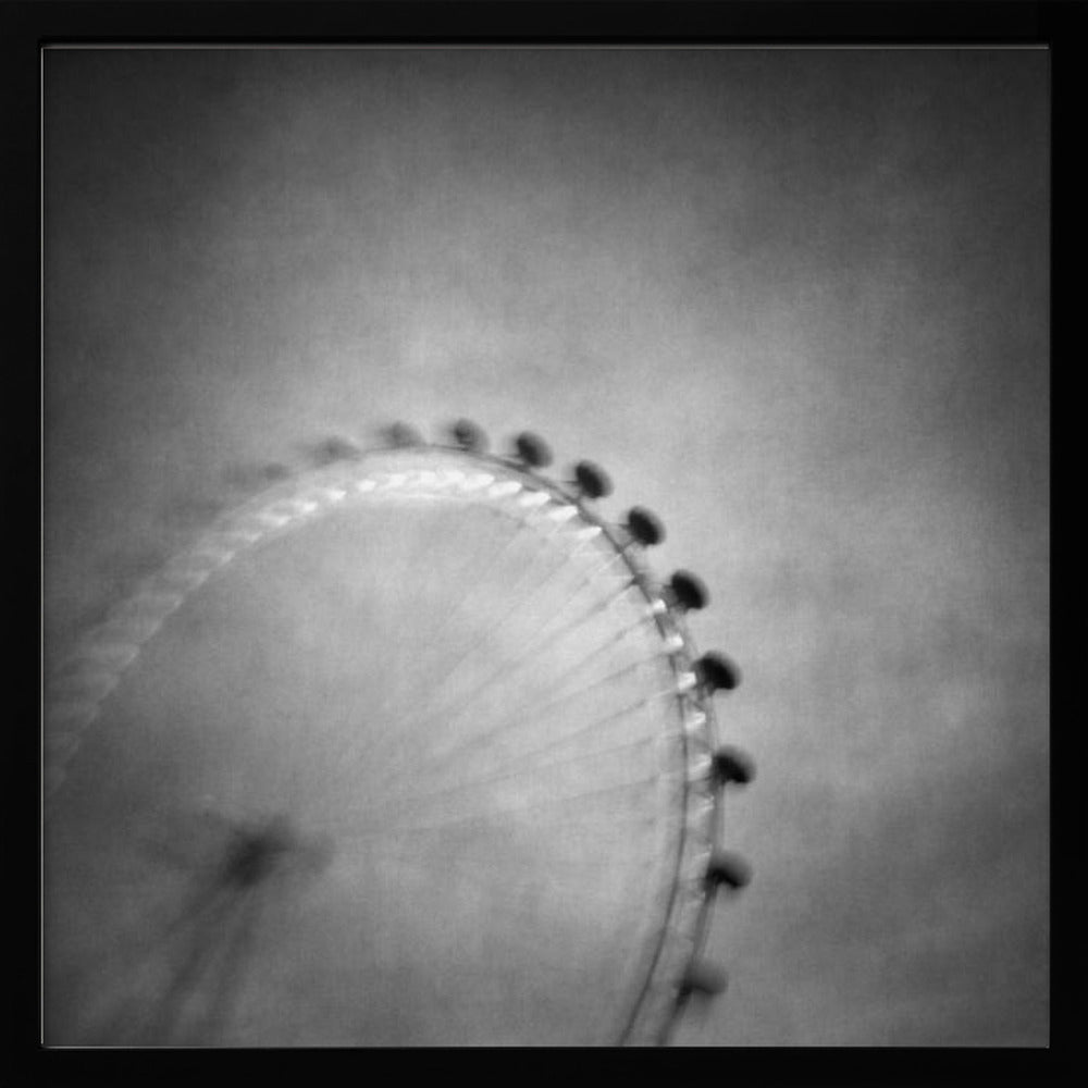 A moody, black and white long-exposure photograph of a large Ferris wheel, creating a soft, blurry effect of its rotation against a grainy gray sky. The artwork is presented in a simple, light wood frame. Poster