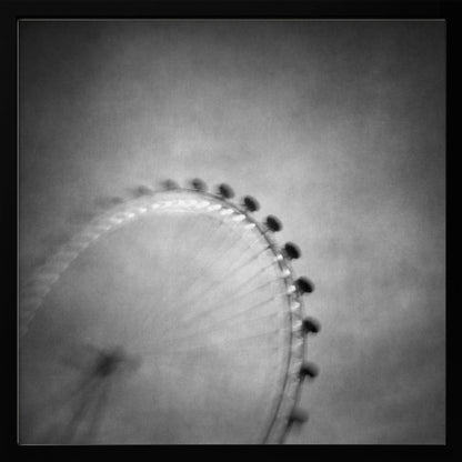 A moody, black and white long-exposure photograph of a large Ferris wheel, creating a soft, blurry effect of its rotation against a grainy gray sky. The artwork is presented in a simple, light wood frame. Poster