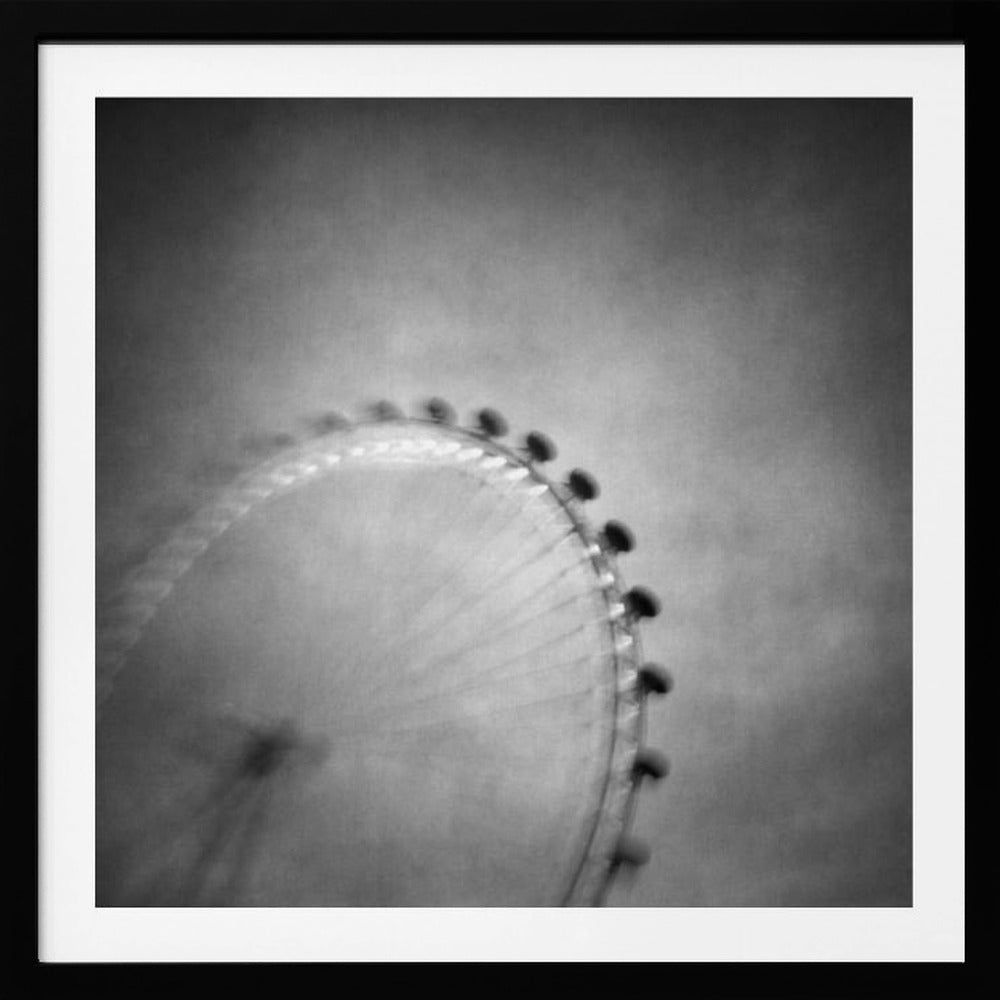 A moody, black and white long-exposure photograph of a large Ferris wheel, creating a soft, blurry effect of its rotation against a grainy gray sky. The artwork is presented in a simple, light wood frame. Poster