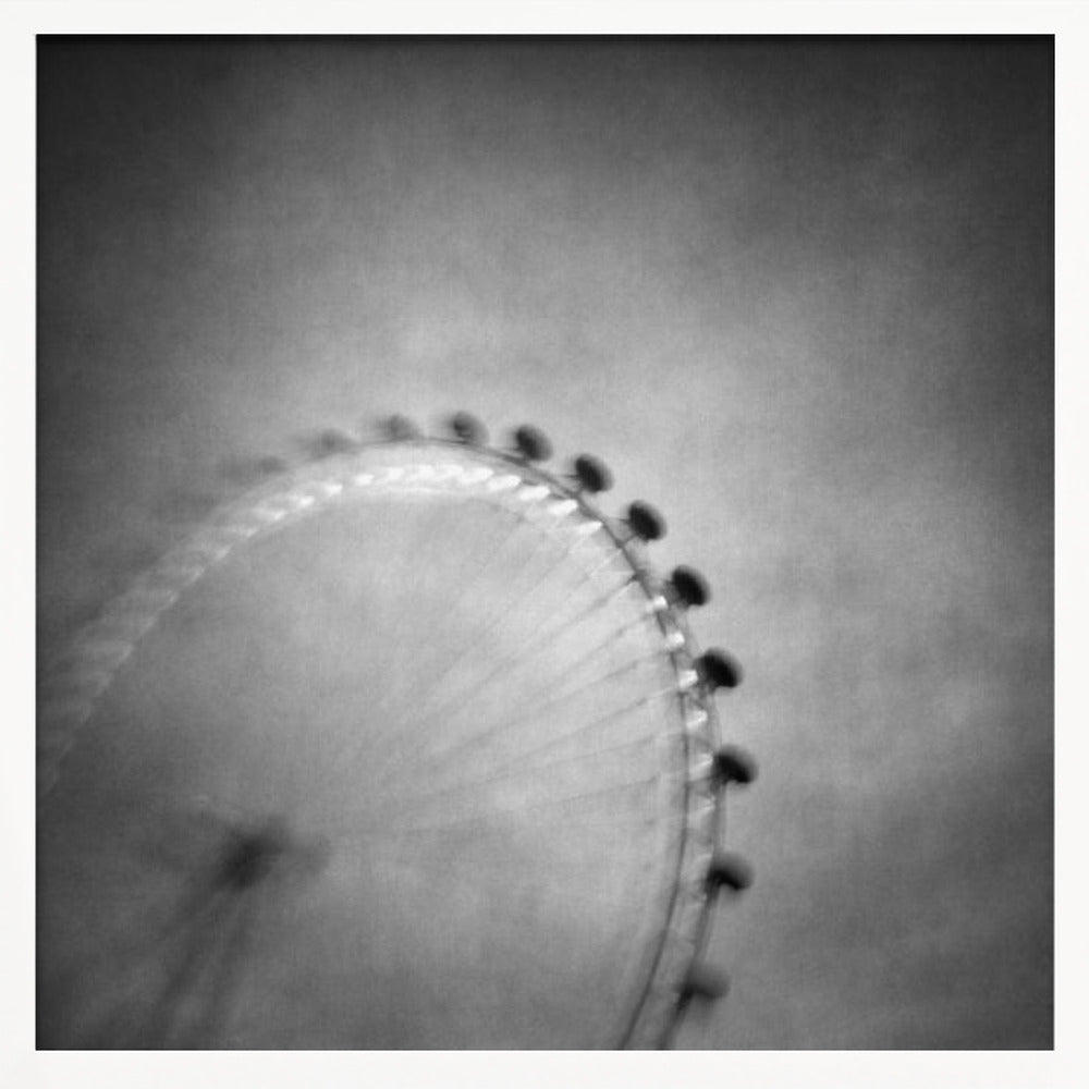 A moody, black and white long-exposure photograph of a large Ferris wheel, creating a soft, blurry effect of its rotation against a grainy gray sky. The artwork is presented in a simple, light wood frame. Poster
