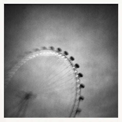 A moody, black and white long-exposure photograph of a large Ferris wheel, creating a soft, blurry effect of its rotation against a grainy gray sky. The artwork is presented in a simple, light wood frame. Poster