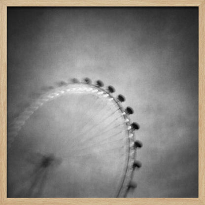 A moody, black and white long-exposure photograph of a large Ferris wheel, creating a soft, blurry effect of its rotation against a grainy gray sky. The artwork is presented in a simple, light wood frame. Poster